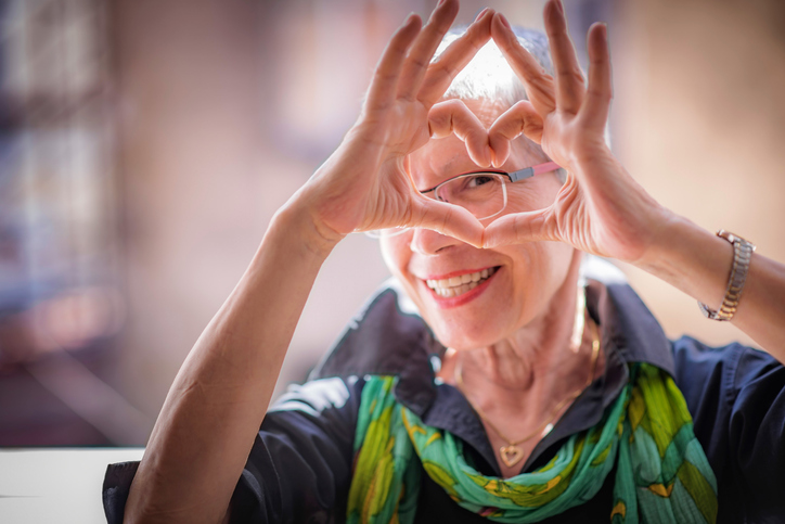 Ältere Dame mit Brille zeigt Herzgeste mit den Fingern vom Gesicht